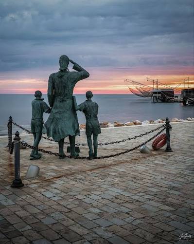 The Sailors' Wives statue in Cesenatico at sunset, with a fishing net platform in the background
